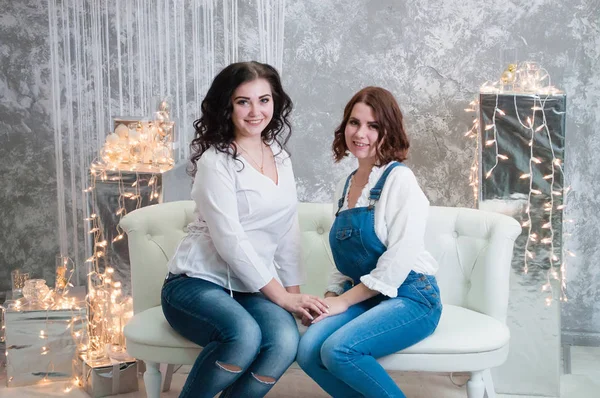 Duas meninas muito bonitas celebram o Natal, ano novo. Meninas no interior do Ano Novo da sala estão sentados em um sofá branco contra o fundo de guirlandas — Fotografia de Stock