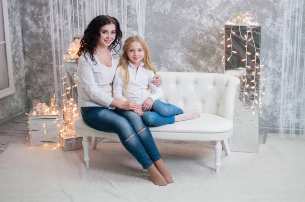 Due ragazze molto carine, le sorelle festeggiano il Natale, il nuovo anno. Le ragazze all'interno della stanza del nuovo anno sono sedute su un divano bianco sullo sfondo delle ghirlande — Foto Stock