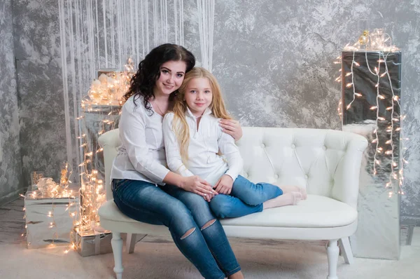 Due ragazze molto carine, le sorelle festeggiano il Natale, il nuovo anno. Le ragazze all'interno della stanza del nuovo anno sono sedute su un divano bianco sullo sfondo delle ghirlande — Foto Stock