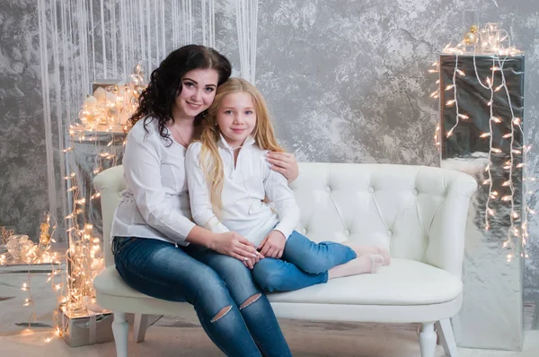 Due ragazze molto carine, le sorelle festeggiano il Natale, il nuovo anno. Le ragazze all'interno della stanza del nuovo anno sono sedute su un divano bianco sullo sfondo delle ghirlande — Foto Stock