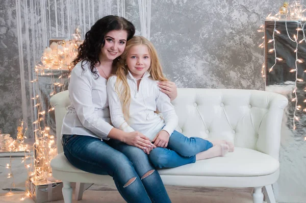 Due ragazze molto carine, le sorelle festeggiano il Natale, il nuovo anno. Le ragazze all'interno della stanza del nuovo anno sono sedute su un divano bianco sullo sfondo delle ghirlande — Foto Stock