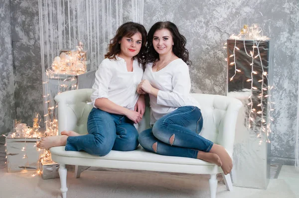 Duas jovens muito bonitas celebram o Natal, o ano novo. Meninas no interior do Ano Novo da sala estão sentados em um sofá branco contra o fundo de guirlandas — Fotografia de Stock