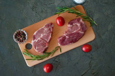 Düz bir zeminde, çiğ et biftekleri, domuz eti, biberiye, deniz tuzu ve domatesler kesme tahtasının üzerinde, koyu gri beton arka planda.