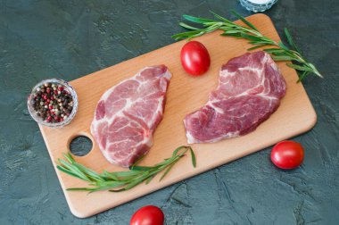 Düz bir zemin, çiğ et, domuz eti, biber, biberiye, deniz tuzu ve domatesler tahtada, koyu gri beton arka planda, yakın plan.