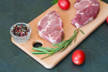 Çiğ biftek, domuz eti, biber, biberiye, deniz tuzu ve domatesler kesme tahtasının üzerinde, koyu gri beton arka planda, yakın plan