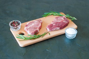 İki çiğ domuz bifteği, deniz tuzu, biberiye ve biber açık ahşap kesim tahtasında, koyu gri beton arka planda.