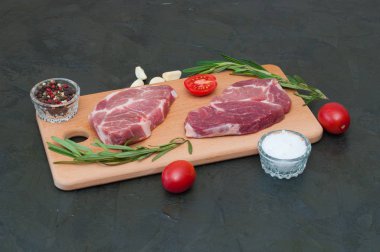 İki çiğ domuz bifteği, deniz tuzu, biberiye, sarımsak, kırmızı domates ve biberler açık ahşap kesim tahtasında, koyu gri beton arka planda.