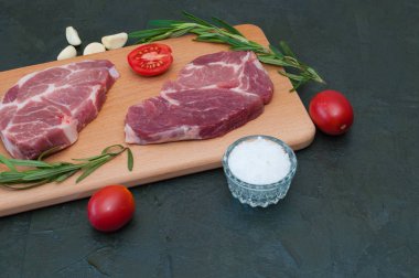 İki çiğ domuz bifteği, deniz tuzu, biberiye, sarımsak, kırmızı domates açık ahşap kesim tahtasının üzerinde, koyu gri beton arka planda fotokopi alanı için sağdan ve alttan alan, üstten görüş.