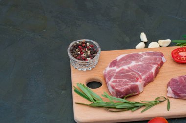 İki çiğ domuz bifteği, deniz tuzu, biberiye, sarımsak, kırmızı domatesler açık ahşap kesim tahtasının üzerinde, koyu gri beton arka planda fotokopi alanı için soldan ve yukarıdan manzara,