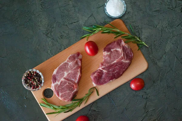 Düz bir zeminde, çiğ et biftekleri, domuz eti, biberiye, deniz tuzu ve domatesler kesme tahtasının üzerinde, koyu gri beton arka planda.