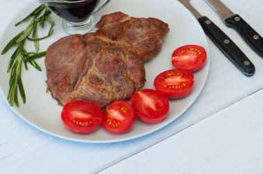 Sos, biberiye ve domatesli gri bir tabakta leziz ızgara domuz bifteği. Hafif ahşap arka planda, çatal bıçak takımında.