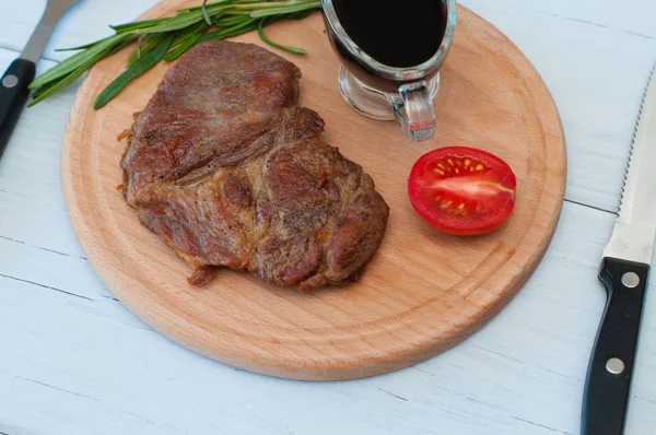 Kızarmış, lezzetli dana eti, biftek ya da domuz eti, domates, biberiye ve soya sosu yuvarlak ahşap bir tahtada, hafif bir arka planda, bıçak ve çatal, yakın plan