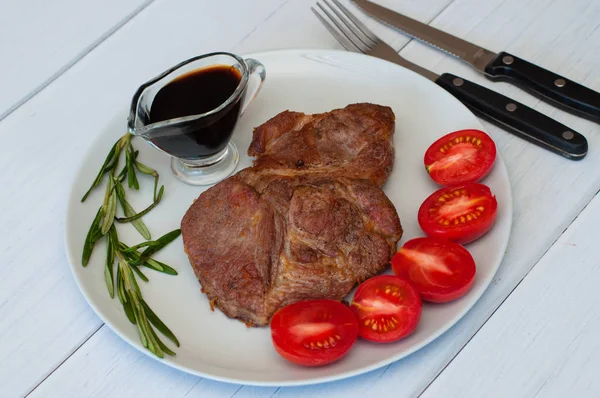 Sos, biberiye ve domatesli gri bir tabakta leziz, ızgara domuz bifteği, hafif ahşap arka plan, çatal bıçak takımı, yakın çekim.