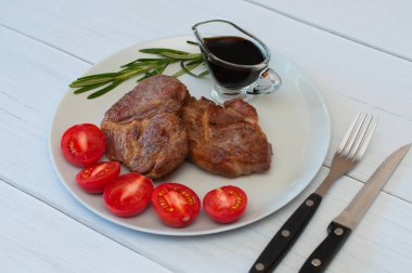 Sos, biberiye ve domatesli gri bir tabakta leziz ızgara domuz bifteği. Hafif ahşap arka planda, çatal bıçak takımında.