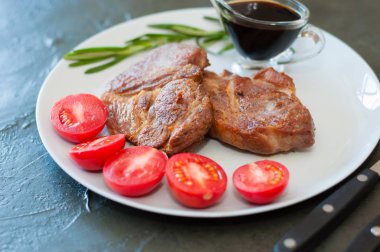 Baharatlı, domatesli ve soslu kokulu domuz eti bifteği gri bir tabakta, metin için yeri olan koyu beton arka planda.