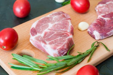 İki çiğ, sulu domuz eti, kırmızı domatesli biftek, biberiye ve sarımsak yakın plan kesim tahtasında.