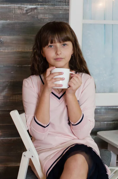 Menina adolescente bonita em um vestido com uma xícara branca de chá na mesa, na varanda da casa. Formato vertical do retrato — Fotografia de Stock