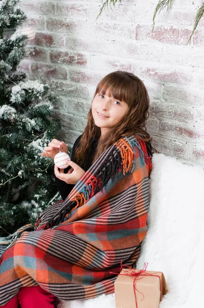 Criança adolescente menina pensativa, sorridente e sonhadora com uma bola de Natal e um presente em um sofá branco perto da árvore de Ano Novo — Fotografia de Stock
