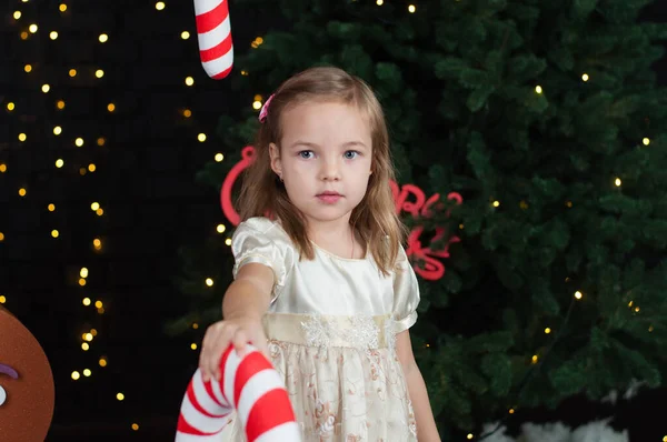 Pequeña chica risueño hermosa en un vestido beige cerca del árbol de Navidad juega con un gran caramelo, sonriendo —  Fotos de Stock
