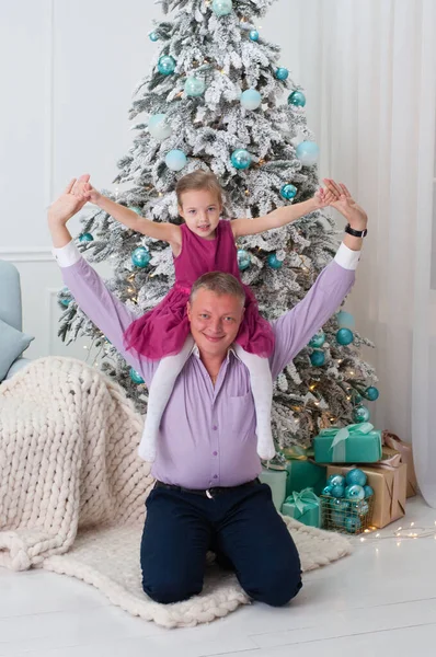 Dad and daughter play merrily, laugh, rejoice around the New Year tree. The child sits on his dad\'s shoulders