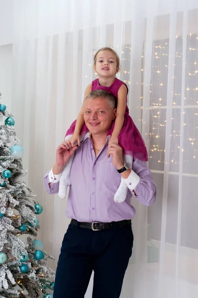 Papà e figlia vicino all'albero di Capodanno in un appartamento o in una casa. Il bambino siede sulle spalle di suo padre — Foto Stock