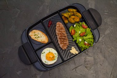 Kahvaltı bifteği ya da pirzola, domatesli ve yeşil sebzeli salata, kızarmış ekmek, patates, yumurta, üstten manzara, koyu arka plan. Sağlıklı beslenme, beslenme, menü, besleyici gıdalar.