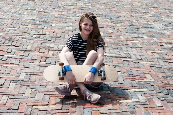 スケートボードを持つかわいい10代の女の子は晴れた夏の日に広場にヨーロッパの都市の中心部の石畳の上に座っています — ストック写真