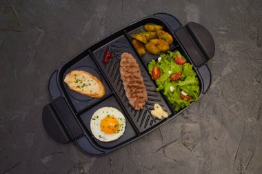 Kahvaltı, kızarmış biftek ya da pirzola, domatesli ve yeşil sebzeli salata, kızarmış ekmek, patates, yumurta, üstten manzara, koyu arkaplan. Sağlıklı beslenme, beslenme, menü, besleyici gıdalar.