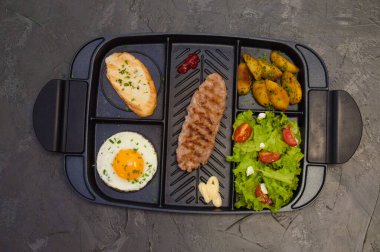 Kahvaltı, biftek ya da pirzola, domatesli ve yeşil sebzeli salata, tost, patates, siyah tabakta yumurta. Sağlıklı beslenme, diyet, menü, iyi beslenme.