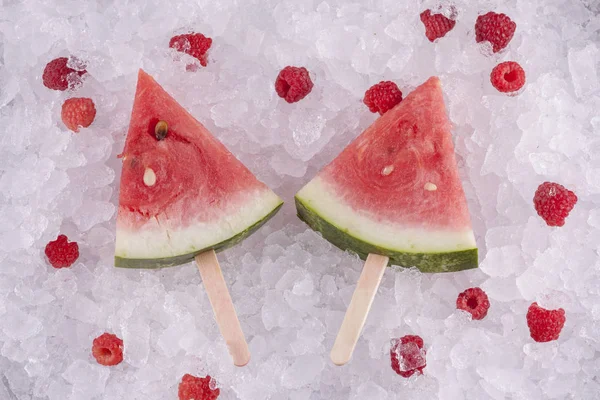 一根冰棒棒上的西瓜片放在冰上 树莓四处散布 — 图库照片