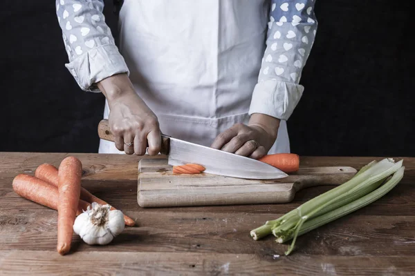 Cutting carrots on a cutting board with celery and garlic on the table.