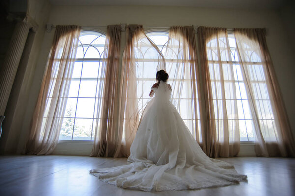 Beautiful bride in long white dress looks out the window.