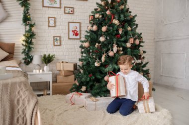 6 yaşında sarışın bir çocuk elinde hediyesiyle bir noel ağacı ve yatağının yanında