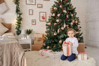 6 yaşında sarışın bir çocuk elinde hediyesiyle bir noel ağacı ve yatağının yanında