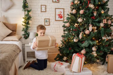6 yaşında sarışın bir çocuk elinde hediyesiyle bir noel ağacı ve yatağının yanında