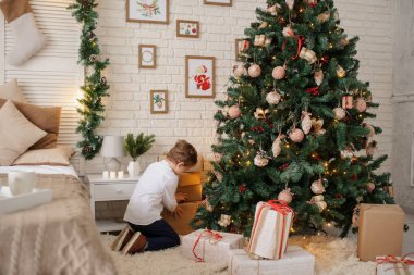 6 yaşında sarışın bir çocuk elinde hediyesiyle bir noel ağacı ve yatağının yanında