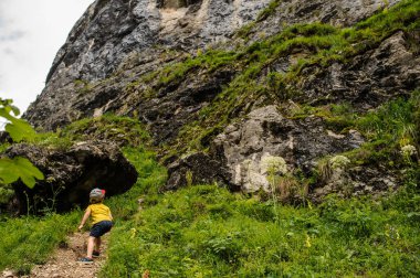 Çimlerle çevrili kayalık bir dağın tepesinde yürüyen 6 yaşında sarışın bir çocuğun sırtı.