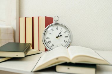 Kitapların yanındaki gece standında gümüş renkli bir saat.