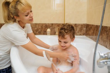 Küçük çocuk annesiyle banyo küvetinde köpük ve sabun köpüğüyle oynuyor.