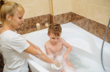Küçük çocuk annesiyle banyo küvetinde köpük ve sabun köpüğüyle oynuyor.