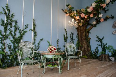 Fotoğraf stüdyosunun iç tasarımı: duvarında akıntılar olan bir ağaç ve iki demir sandalye ve üzerinde kırmızı ve beyaz güller ve östoma olan bir gelin buketi olan bir masa.