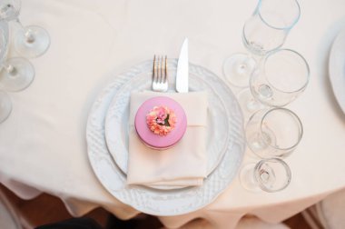 Katlanmış bir peçetedeki çatal ve bıçağın üst görüntüsü. Bardakların yanında iki tabakta ve peçetenin üzerinde çiçek olan pembe yuvarlak bir hediye kutusu.