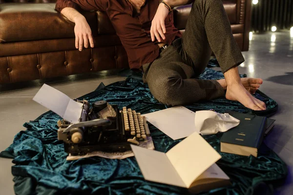 lateral view of man sitting near a typewriter on a blue velvet fabric on the floor in a dark room with light bulbs near a brown leather sofa with sheets of paper and books arround