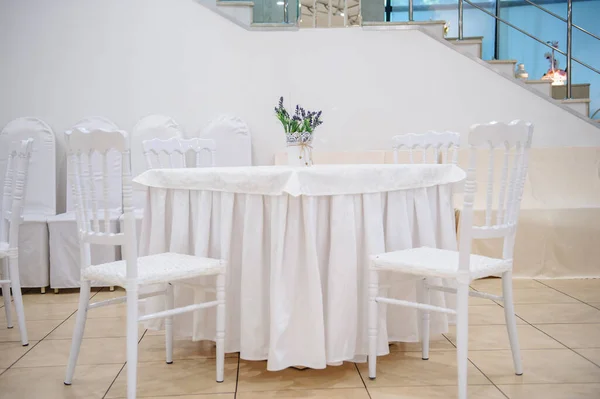 lateral view of a square table with white tablecloth and a little vase with flower on it surrounded by four white chairs in a banquet hall