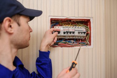 lateral view of a young eletrician in blue overall disassembling a electrical panel with fuses in a hous clipart