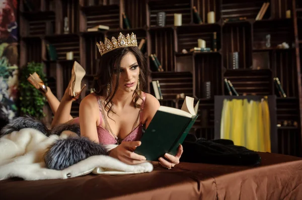 close up photo of a sexy woman laying on a white fur coat with gray collar, in pink underwear with a crown on her head, reading a book near bookshelves with books