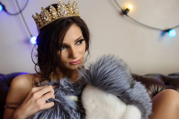 close up photo of a sexy woman in white fur coat with gray collar, in pink underwear with a crown laying on a brown leather sofa near a wall with colorful light bulbs