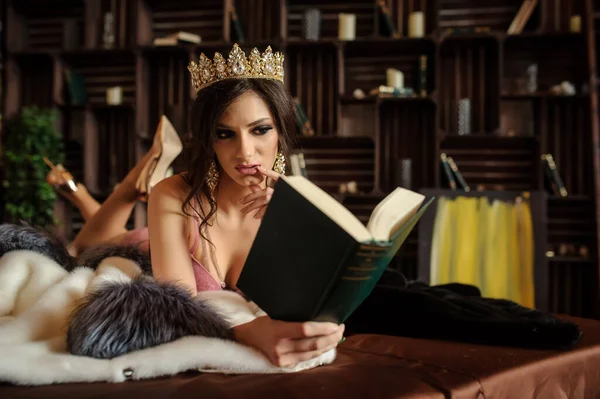 close up photo of a sexy woman laying on a white fur coat with gray collar, in pink underwear with a crown on her head, reading a book near bookshelves with books