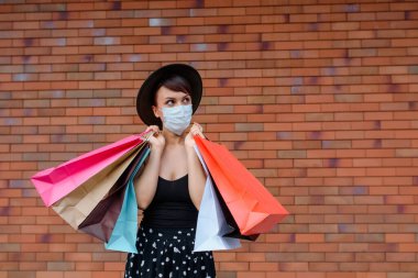 Koruyucu maskeli genç bir kadın güzel bir alışveriş gününden sonra bir sürü renkli alışveriş torbası tutuyor. Coronavirus covid-19 pandemisi. Metin için boşluk kopyala