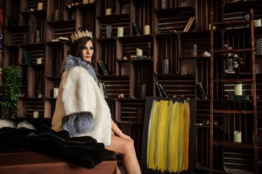 Kafasında taç olan pembe iç çamaşırlı, gri yakalı beyaz kürklü, kitaplarla kitaplıkların yanındaki bir yatakta oturan bir kadının yan görüntüsü.